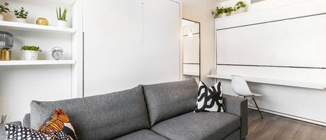 A modern living room with a grey sofa, a white coffee table, and a white chair. There are shelves with plants and decorative items.