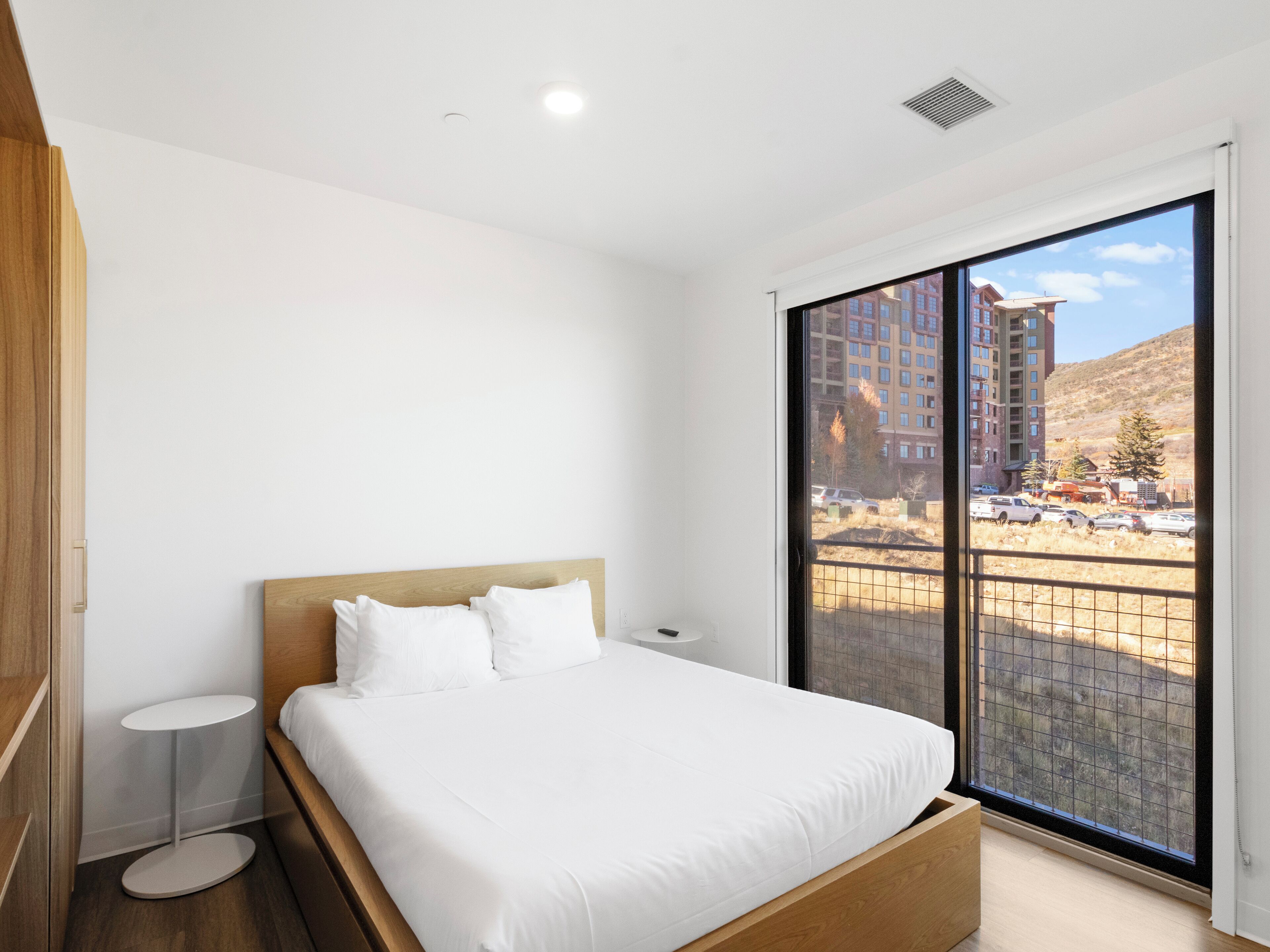 A bedroom with a wooden bed, white bedding, a small bedside table, and a large window with a view of buildings and vehicles outside.