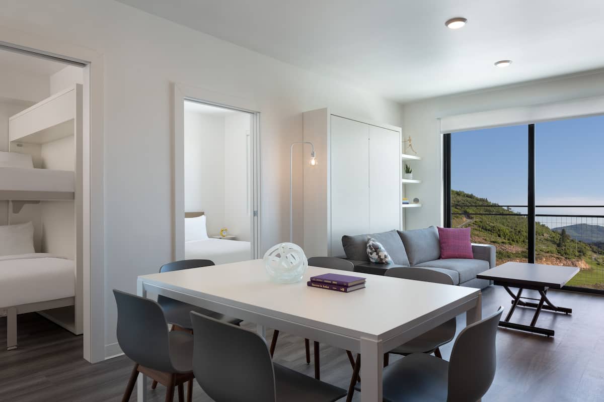 Un salon moderne avec une table à manger blanche, des chaises grises, un canapé avec des coussins et une vue sur l’extérieur grâce à de grandes fenêtres.
