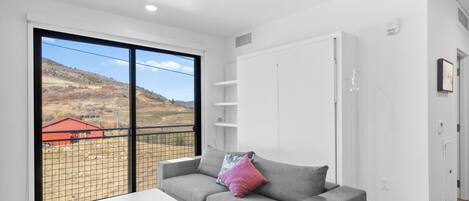 Sala moderna com sofá cinzento, mesa de centro branca e uma grande janela com vista para um edifício vermelho e colinas.
