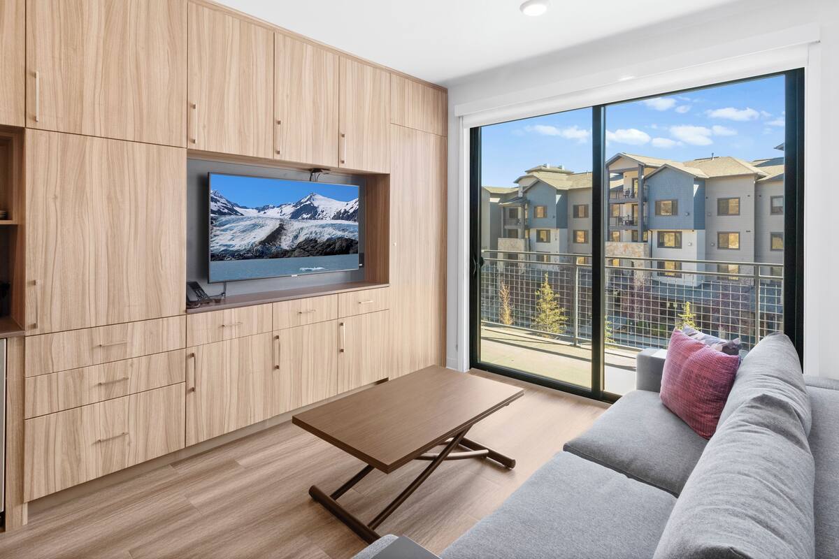 Un salon moderne avec un téléviseur à écran plat fixé à un mur en bois, un canapé gris agrémenté d’un coussin rouge et une table basse en bois.