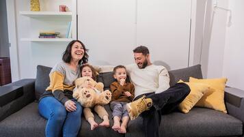 A family of four on a grey sofa with a yellow pillow, a bookshelf in the background, and a white wall.