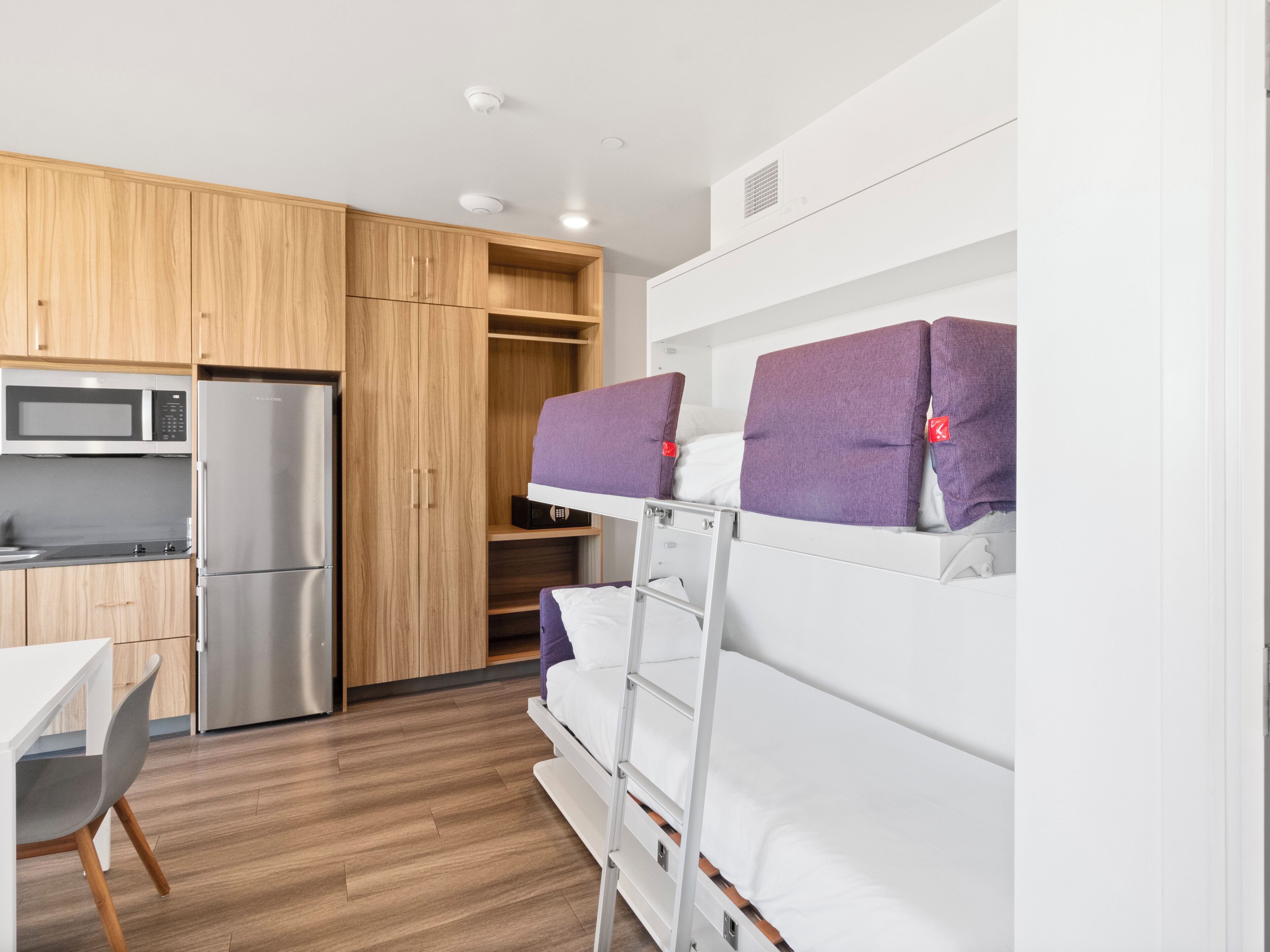 A compact kitchen with a microwave, refrigerator, and wooden cabinets. There is a bunk bed with purple pillows and a white table with a chair.