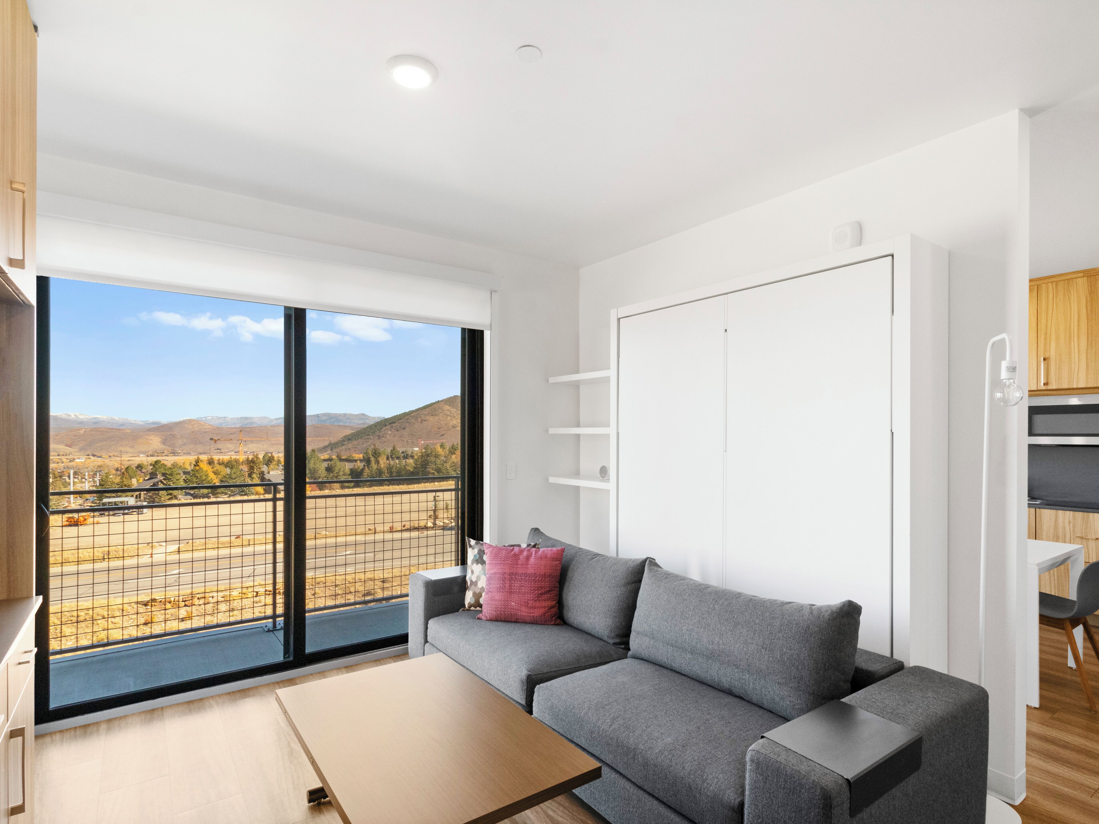 A modern living room with a grey sofa, a wooden coffee table, and a large window with a view of a mountainous landscape.