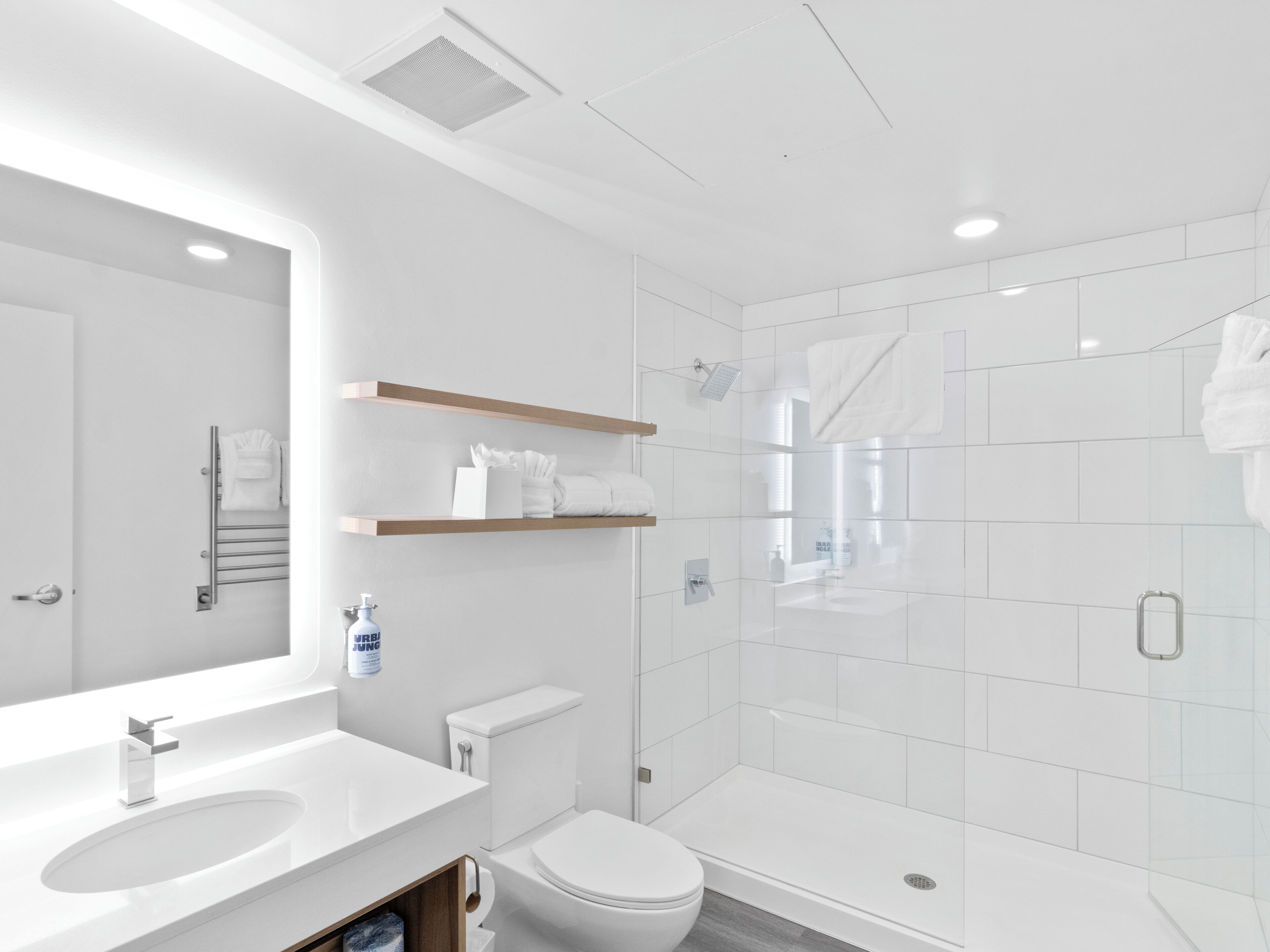 A modern bathroom with a white sink, toilet, and shower. There are wooden shelves above the toilet and a mirror above the sink.