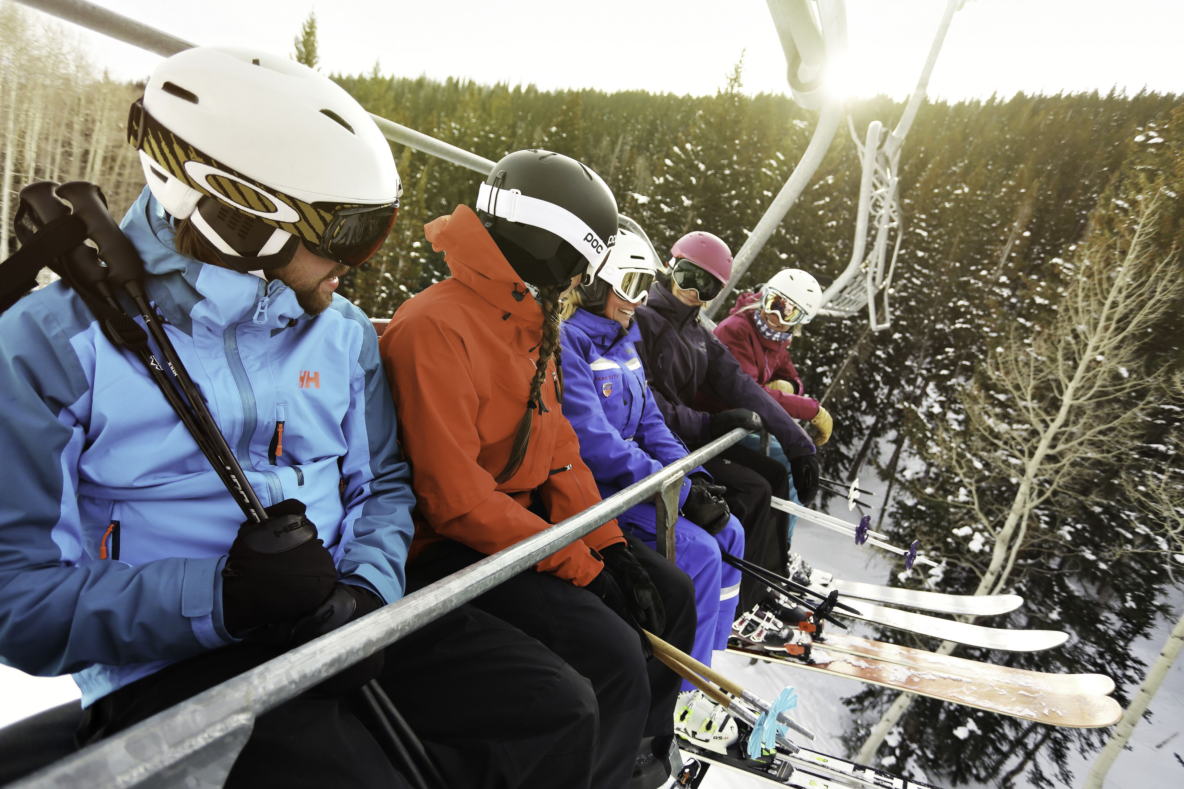 A group of people seated on a ski lift, wearing helmets and ski gear, with snow-covered trees in the background.
