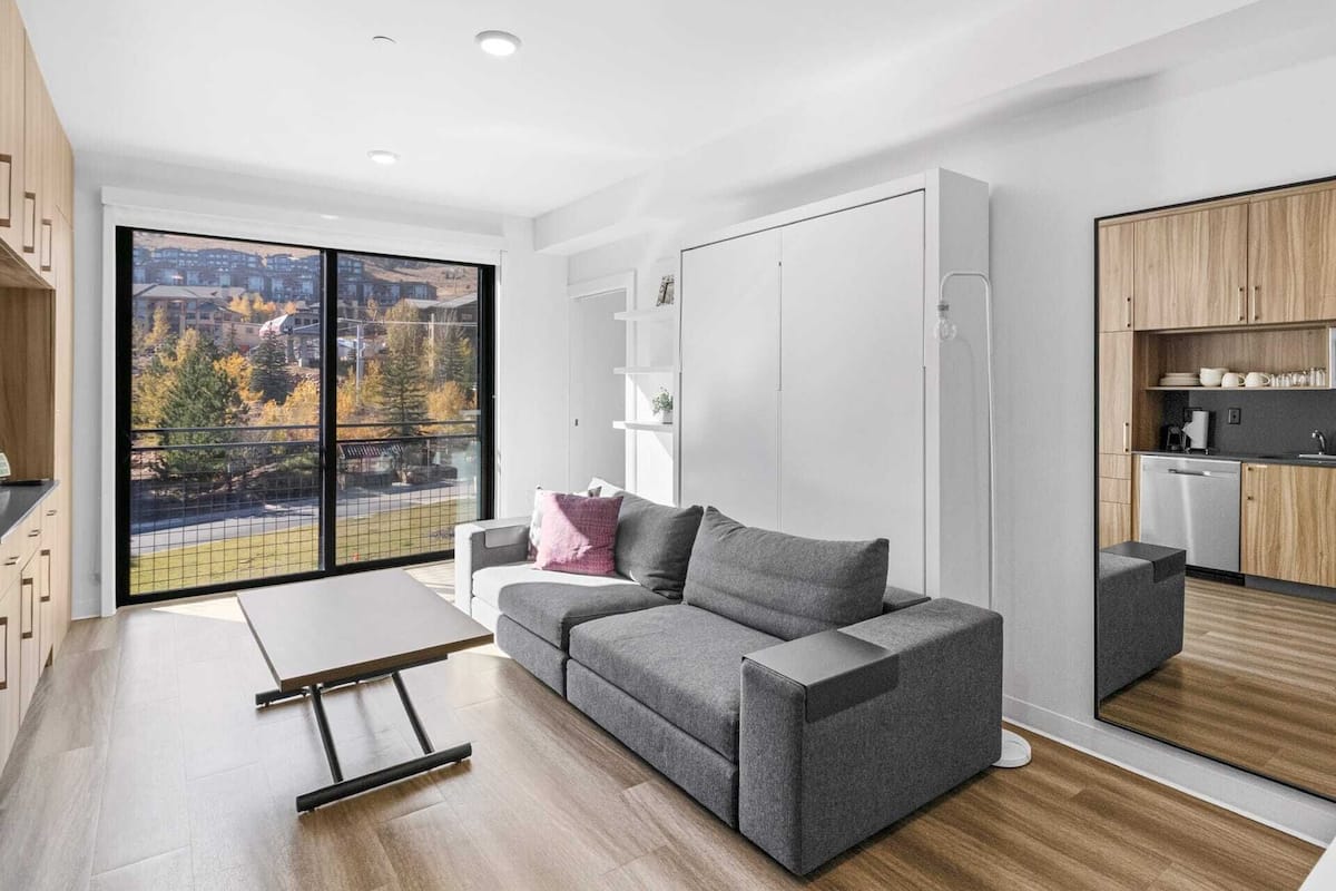 Un salon moderne avec un canapé gris, une petite table et une vue sur le paysage urbain, visible à travers de grandes fenêtres.