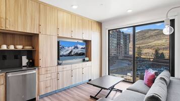 A modern kitchen with wooden cabinets, a built-in TV, and a dining area with a grey sofa and a table.