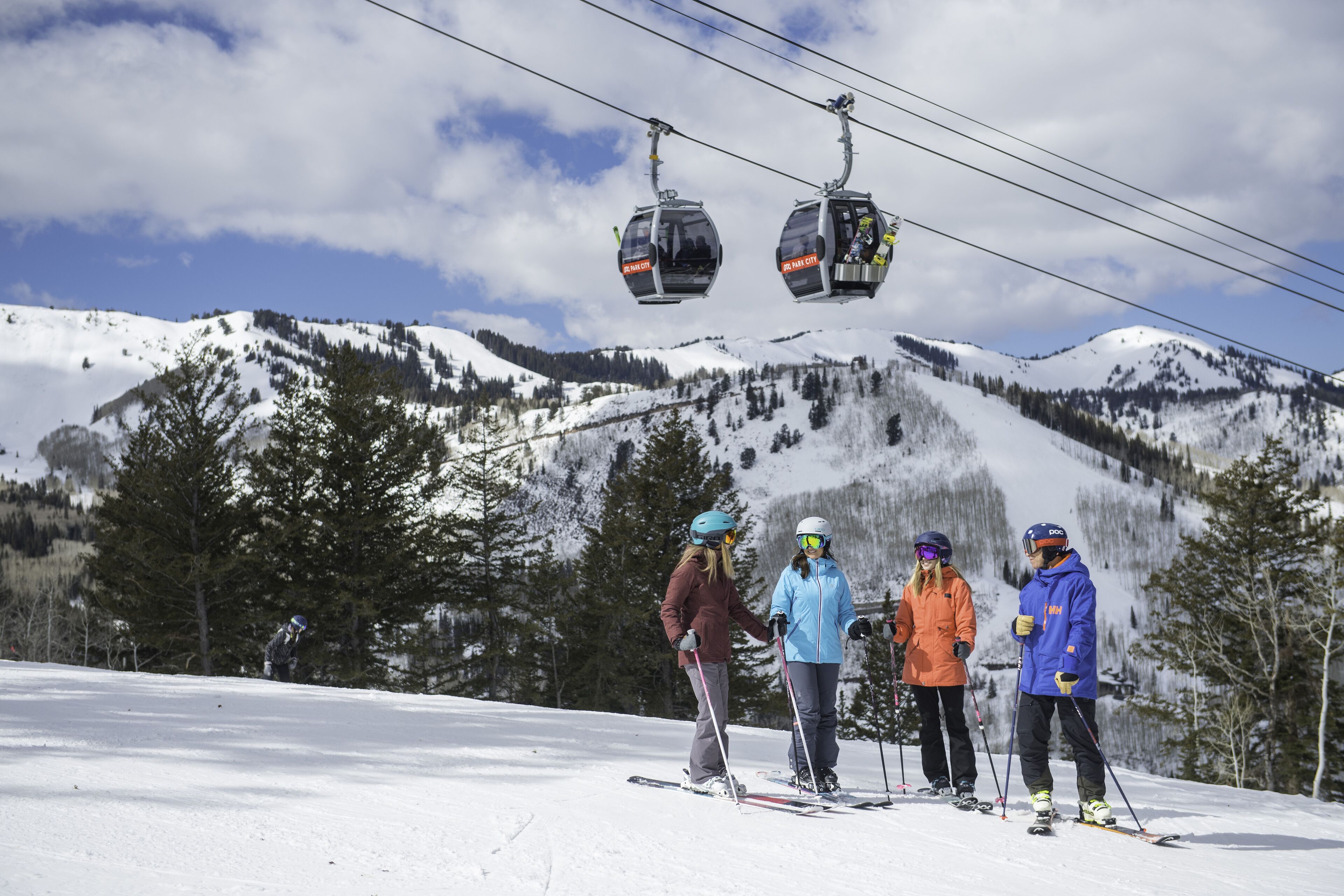A ski lift with four chairs, a snowy mountain backdrop, and skiers on the slope.