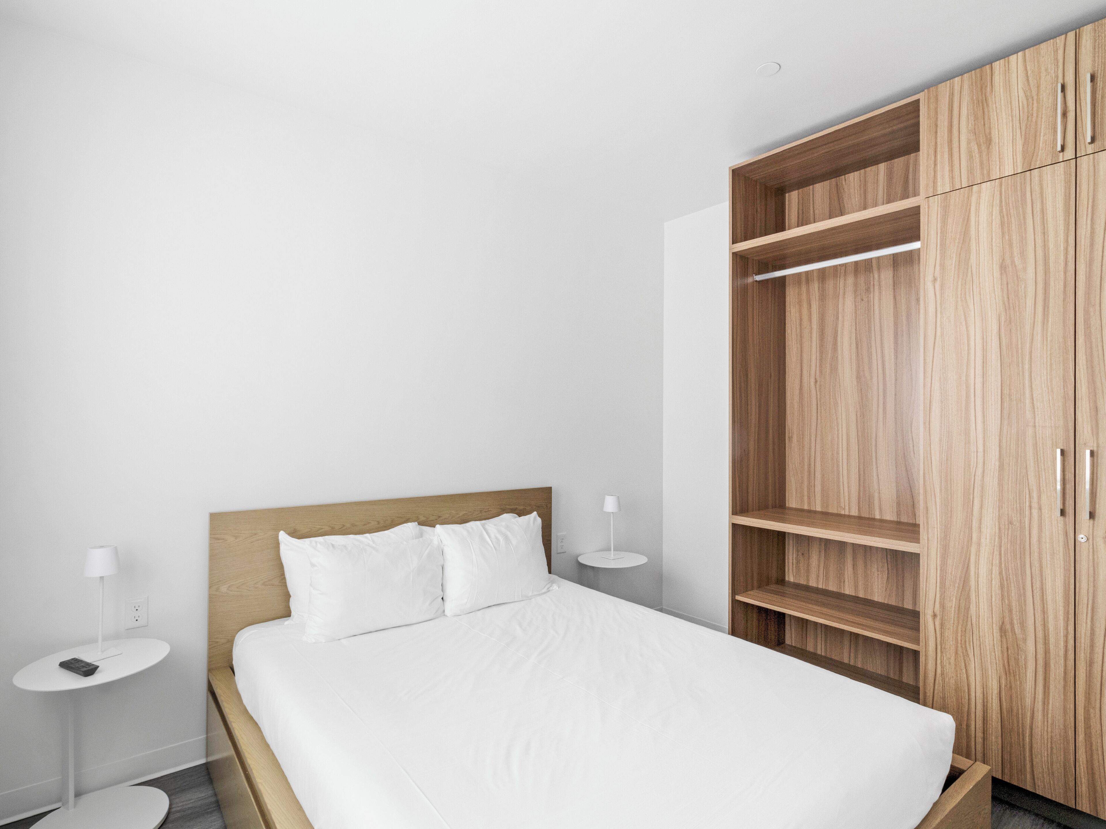 Een moderne slaapkamer met een houten bedframe, wit beddengoed en een grote houten garderobe met veel planken.