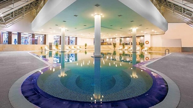 A large indoor swimming pool with a central column, surrounded by a tiled area and a high ceiling with visible structural beams. - Ramada Plaza Hotel (Astana)