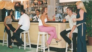 A bar area with stools and a bar counter, shelves with bottles, and a potted plant. - Palmin Hotel (Kuşadası)