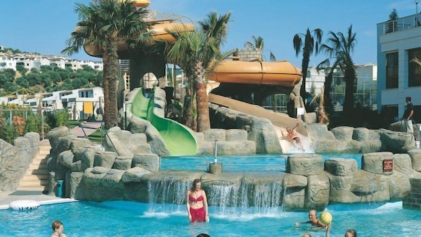 A pool area with water slides, palm trees, and a waterfall feature. - Palmin Hotel (Kuşadası)