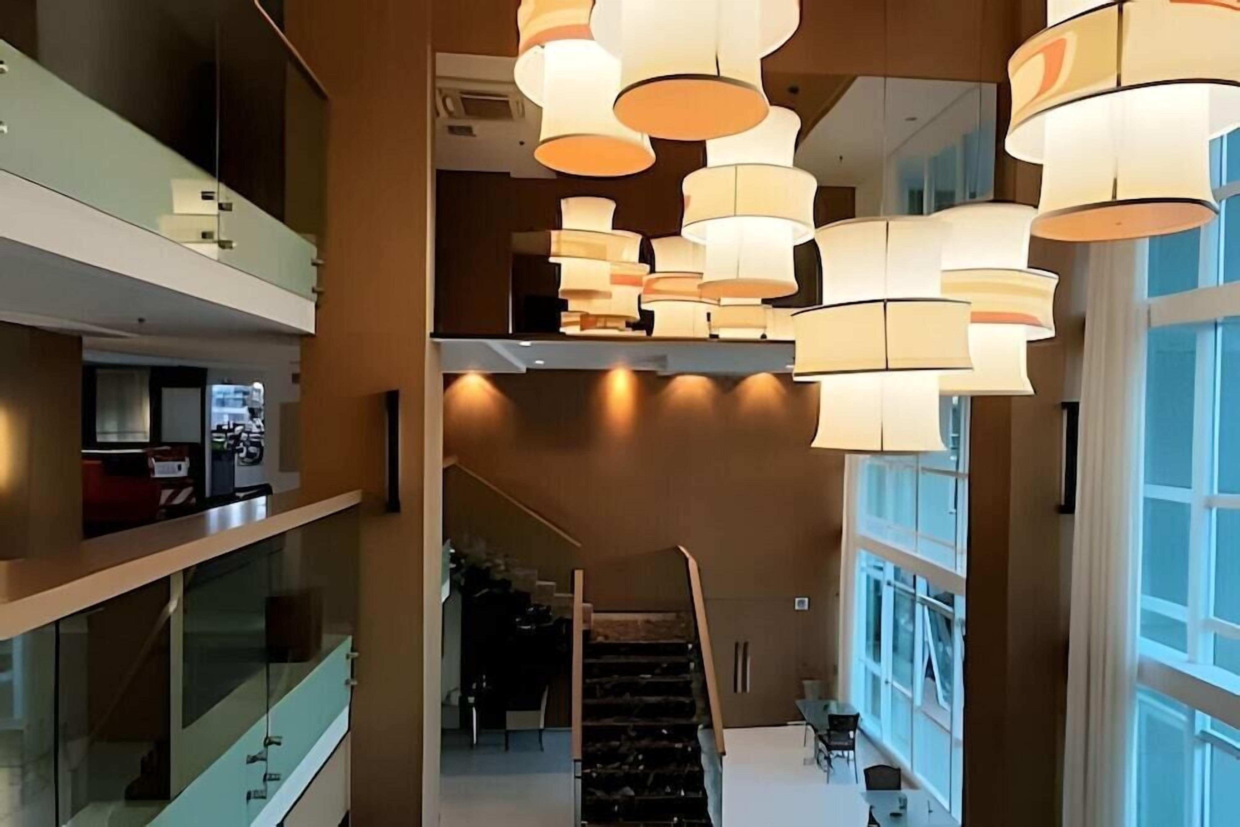 Um hall de hotel moderno com escadaria, grandes janelas e várias luminárias suspensas.