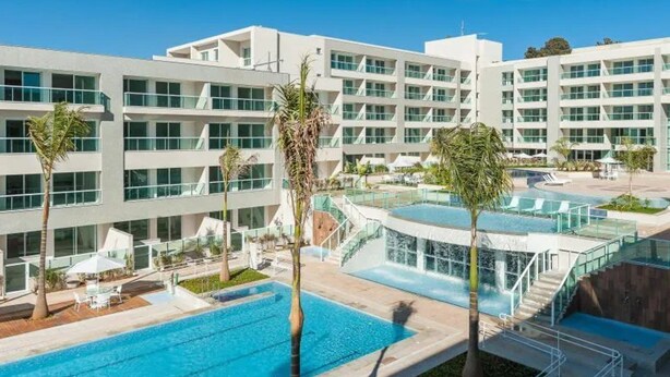 Een modern hotel met een centraal gelegen zwembad, meerdere balkons en een verzorgd buitengebied met palmbomen.