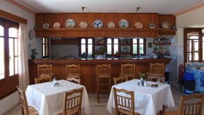 A dining area with wooden tables and chairs, a bar with shelves of bottles, and a kitchen area with cabinets and windows. - Anemoessa (Karpathos)