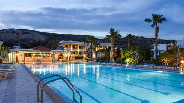 A swimming pool with a tiled deck, a bar area, and a view of buildings and hills in the background. - Chrysoula (Kos)