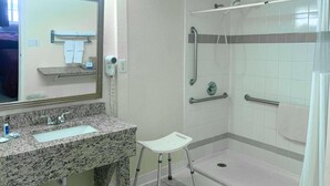 A bathroom with a granite countertop sink, a mirror, a white chair, and a walk-in shower with grab bars.