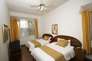 A hotel room with two beds, a ceiling fan, a window with curtains, and a painting on the wall. - 1492 Hotel (San José)
