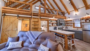 Ein großzügiger Loft-Raum mit hohen Decken, freiliegenden Holzbalken, einem gemütlichen Ledersofa mit Kissen, einem Holzessttisch und einer Küchenzeile mit modernen Geräten.