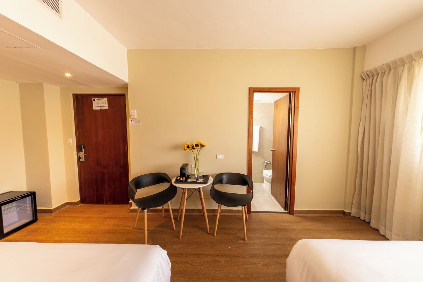 Une chambre d’hôtel avec deux lits, une télévision, une petite table sur laquelle est posé un vase rempli de tournesols, et une porte donnant sur une salle de bain.