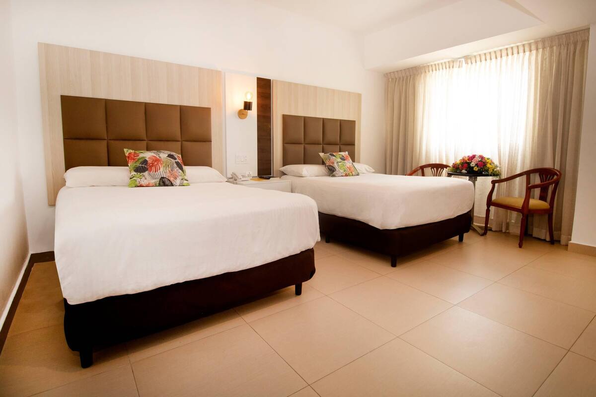 A hotel room with two beds, a chair, a small table with a vase of flowers, and a window with curtains.