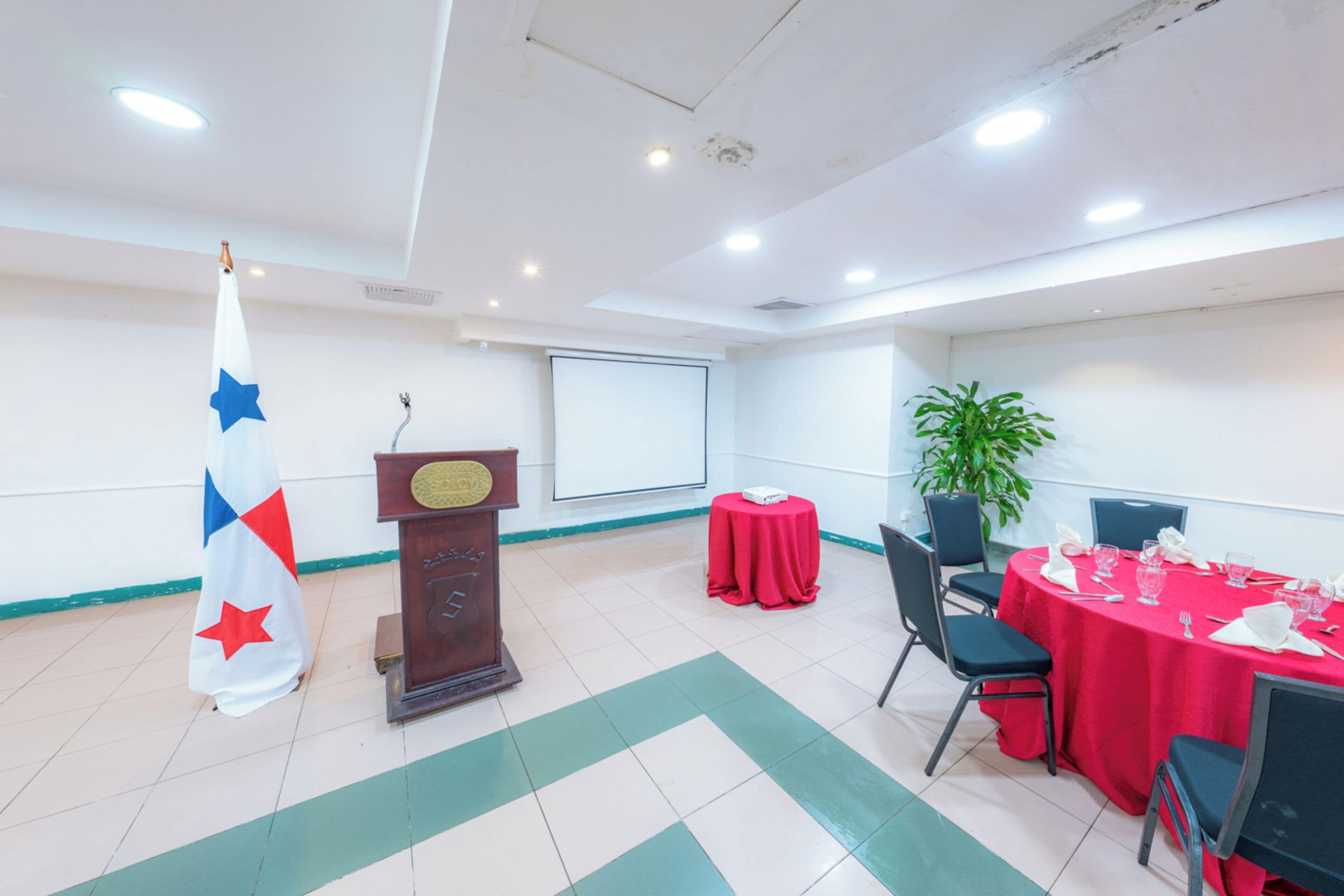 Un salón de conferencias con un atril, una pantalla de proyector, una bandera, una mesa con sillas y una planta en maceta.