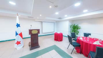 Un salón de conferencias con un atril, una pantalla de proyector, una bandera, una mesa con sillas y una planta en maceta.