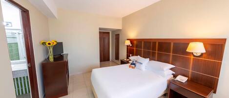 Une chambre d’hôtel avec un grand lit, une table de chevet avec une lampe, une tête de lit en bois et un bureau avec un écran d’ordinateur et un vase rempli de tournesols.