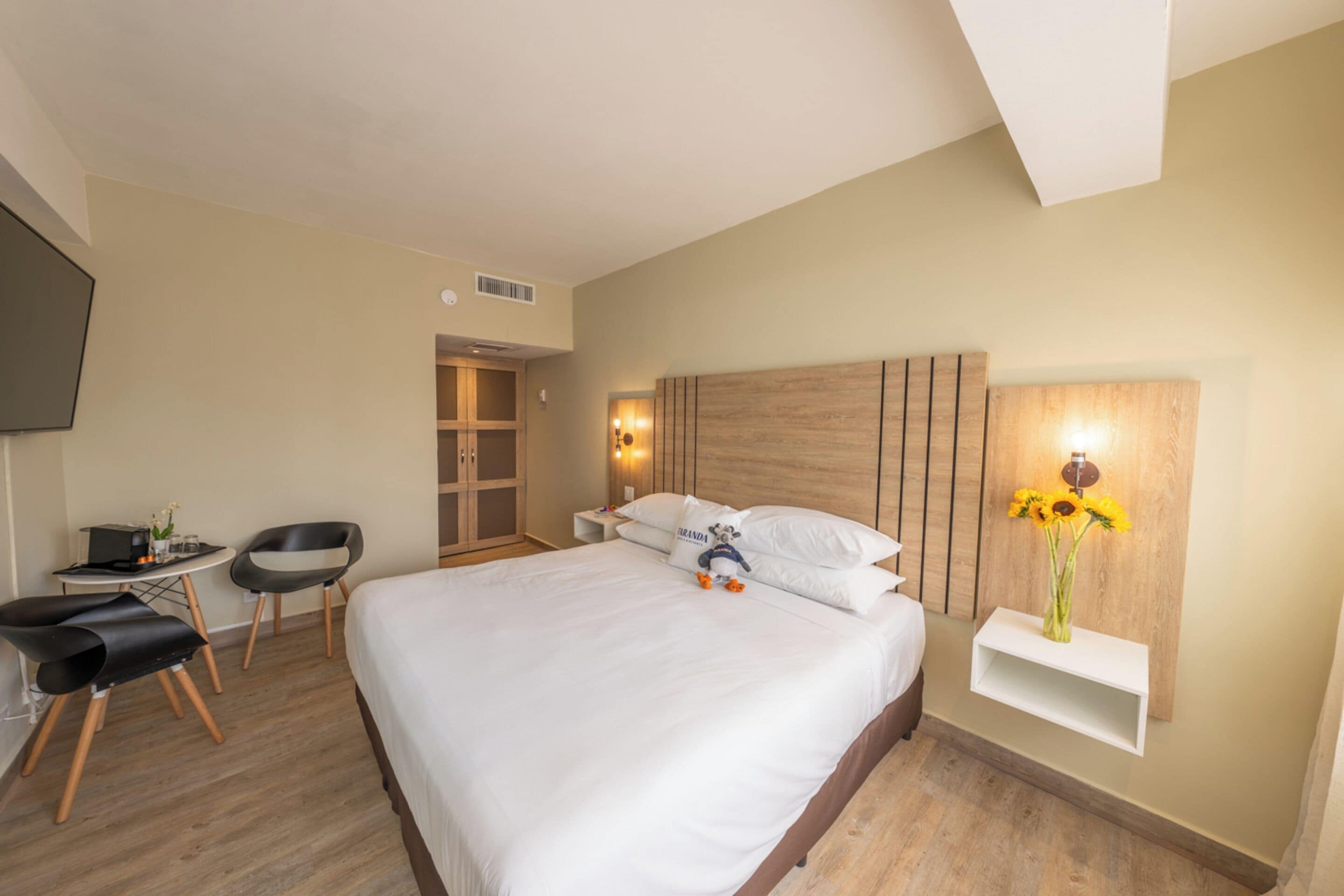 A hotel room with a large bed, a desk with chairs, a wardrobe, and a wall-mounted shelf with flowers.