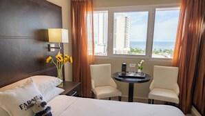 Habitación de hotel con cama, dos sillones, una mesita de noche con lámpara, un jarrón con girasoles y vistas a edificios altos y al mar.