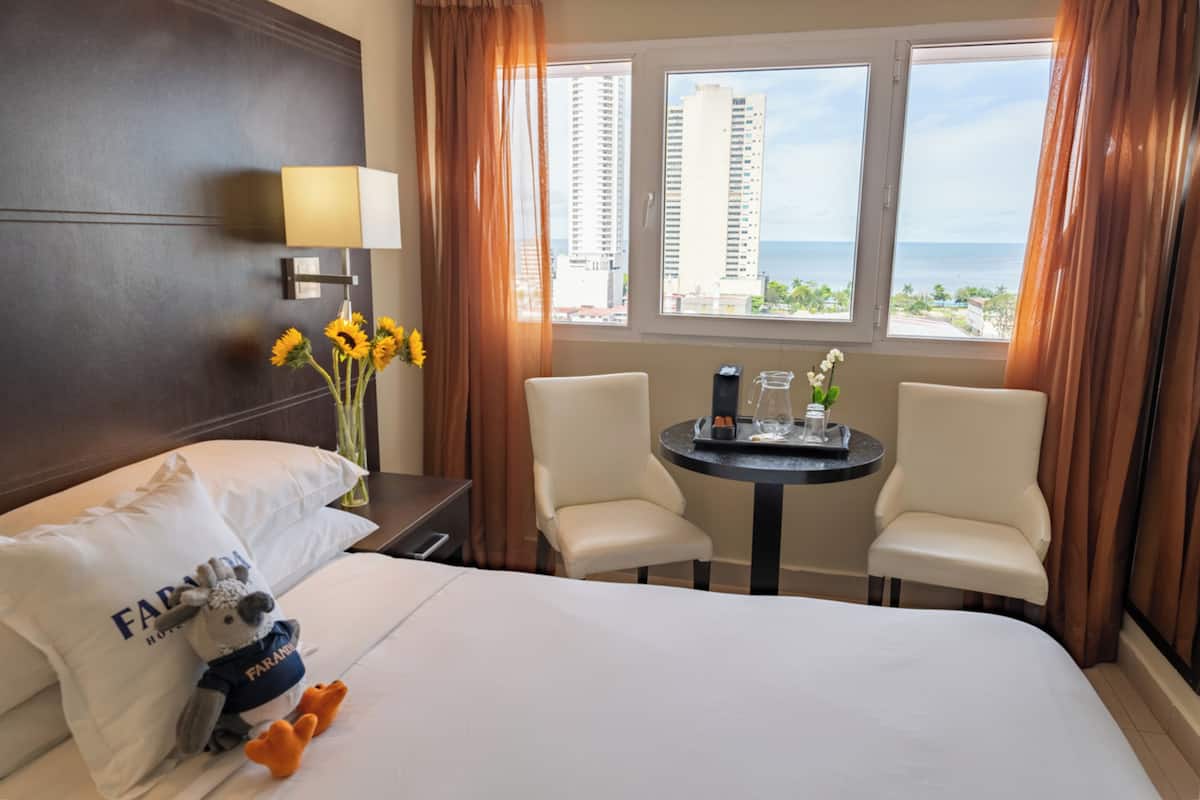 A hotel room with a bed, two chairs, a small table with a lamp, a vase with sunflowers, and a view of tall buildings and the sea.