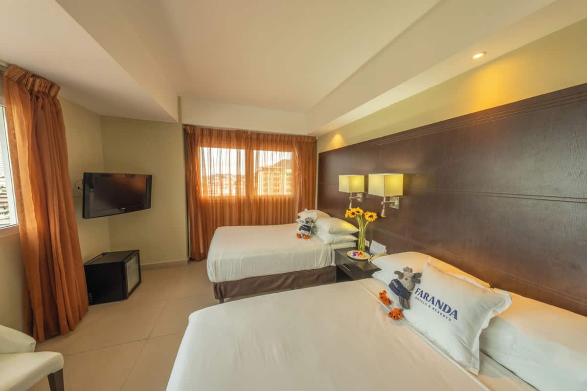A hotel room with two beds, a flat-screen TV, a desk with a lamp, and a view of the city.