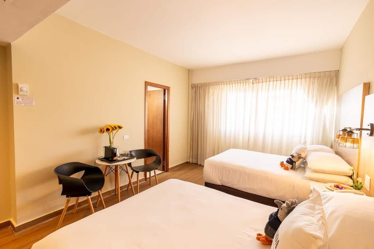A hotel room with two beds, a small table with chairs, a vase of sunflowers, and a dog on the bed.