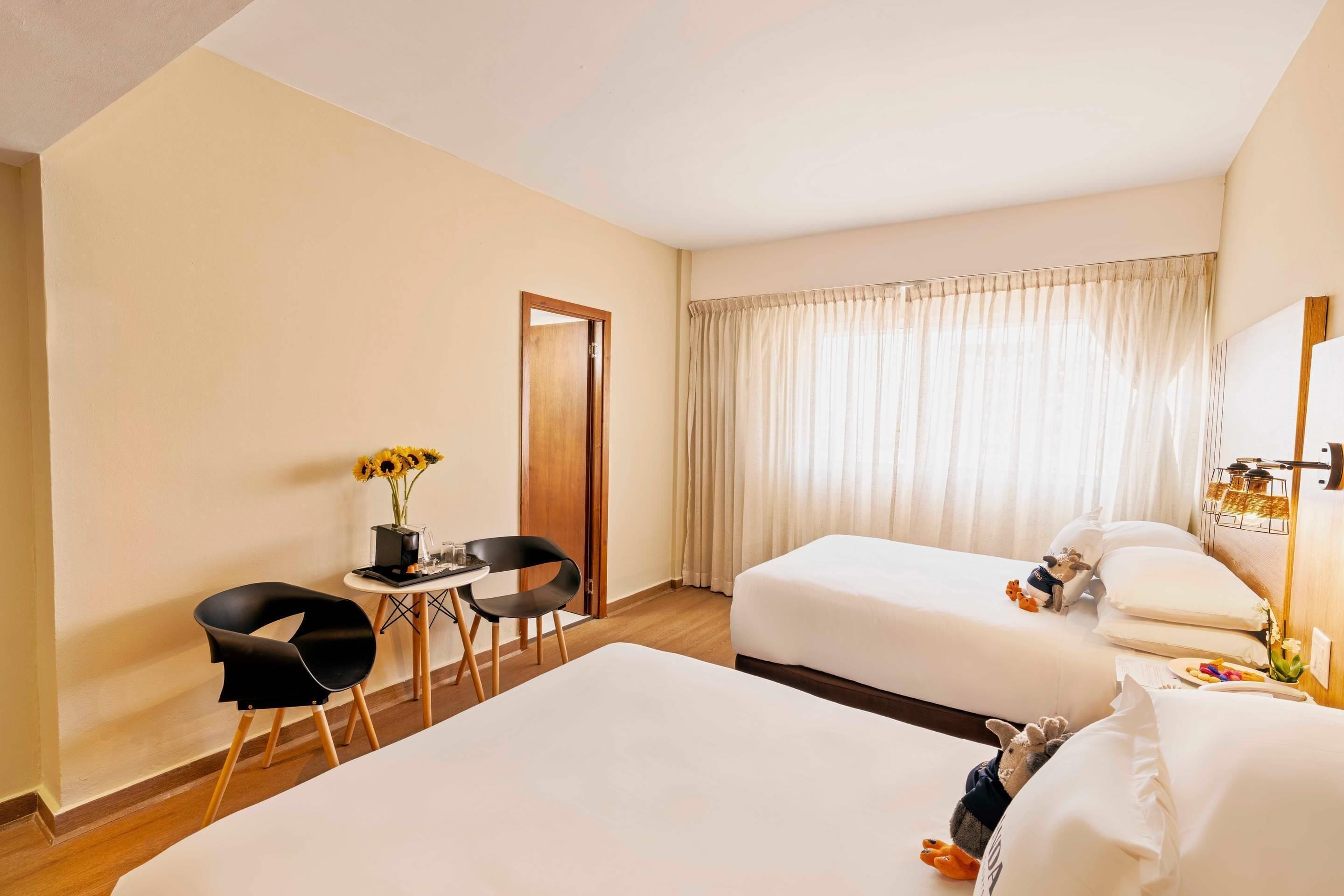 Quarto de hotel com duas camas, uma mesa com cadeira, uma janela com cortinas e uma pequena mesa com um vaso de girassóis.