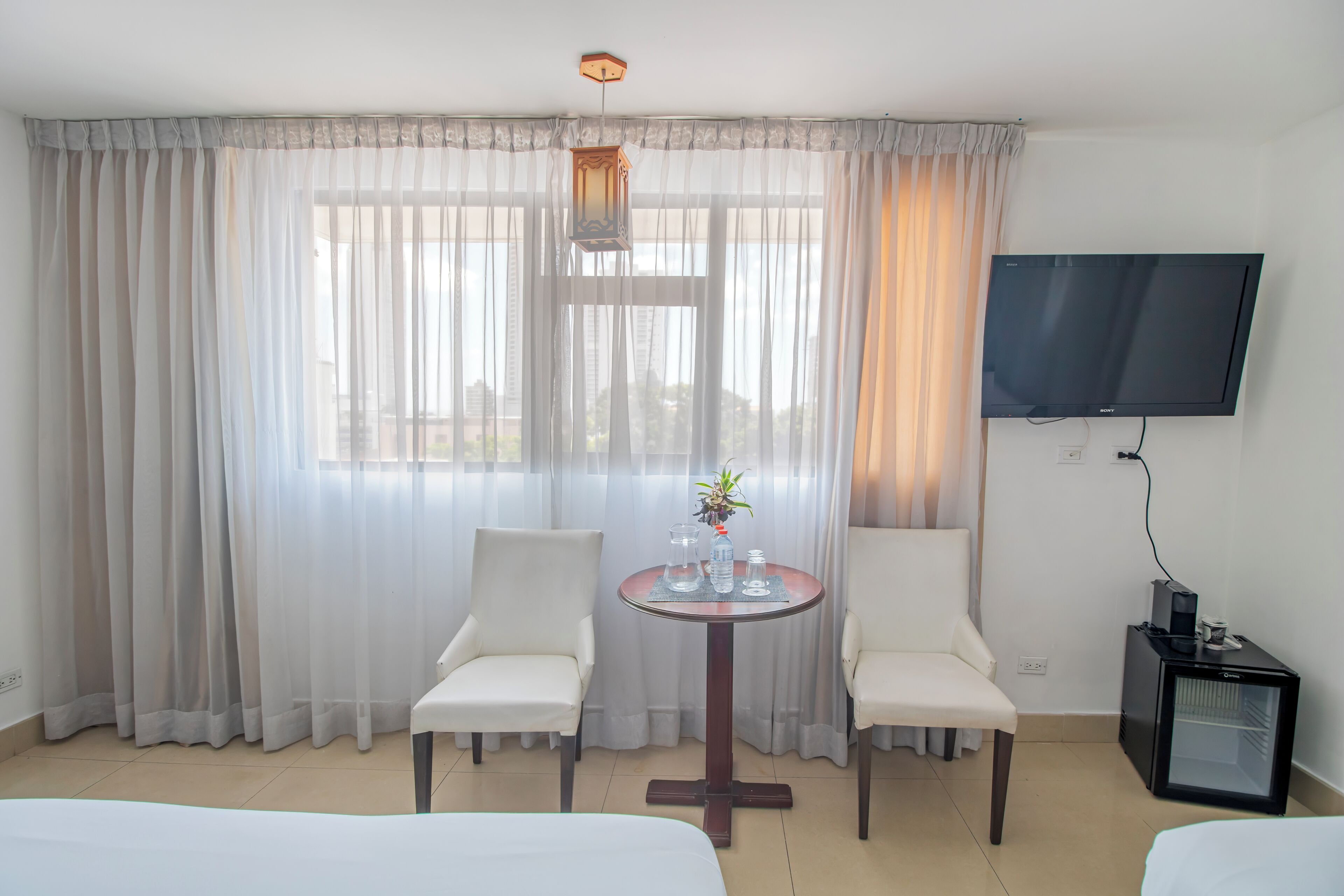 A hotel room with two white chairs, a round table with a plant, a TV, and a mini-fridge.