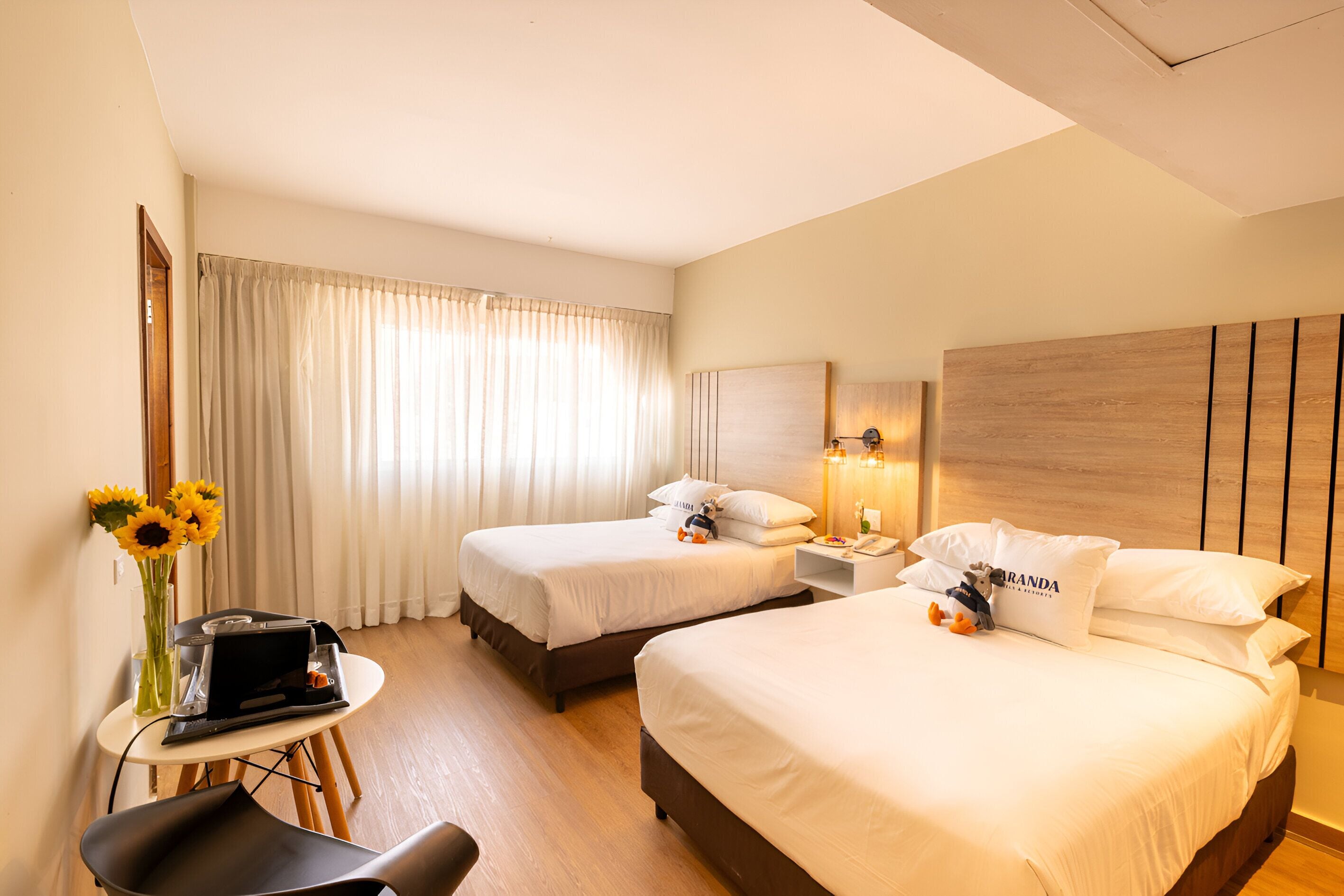 A hotel room with two beds, a desk with a laptop, a chair, and a vase with sunflowers.