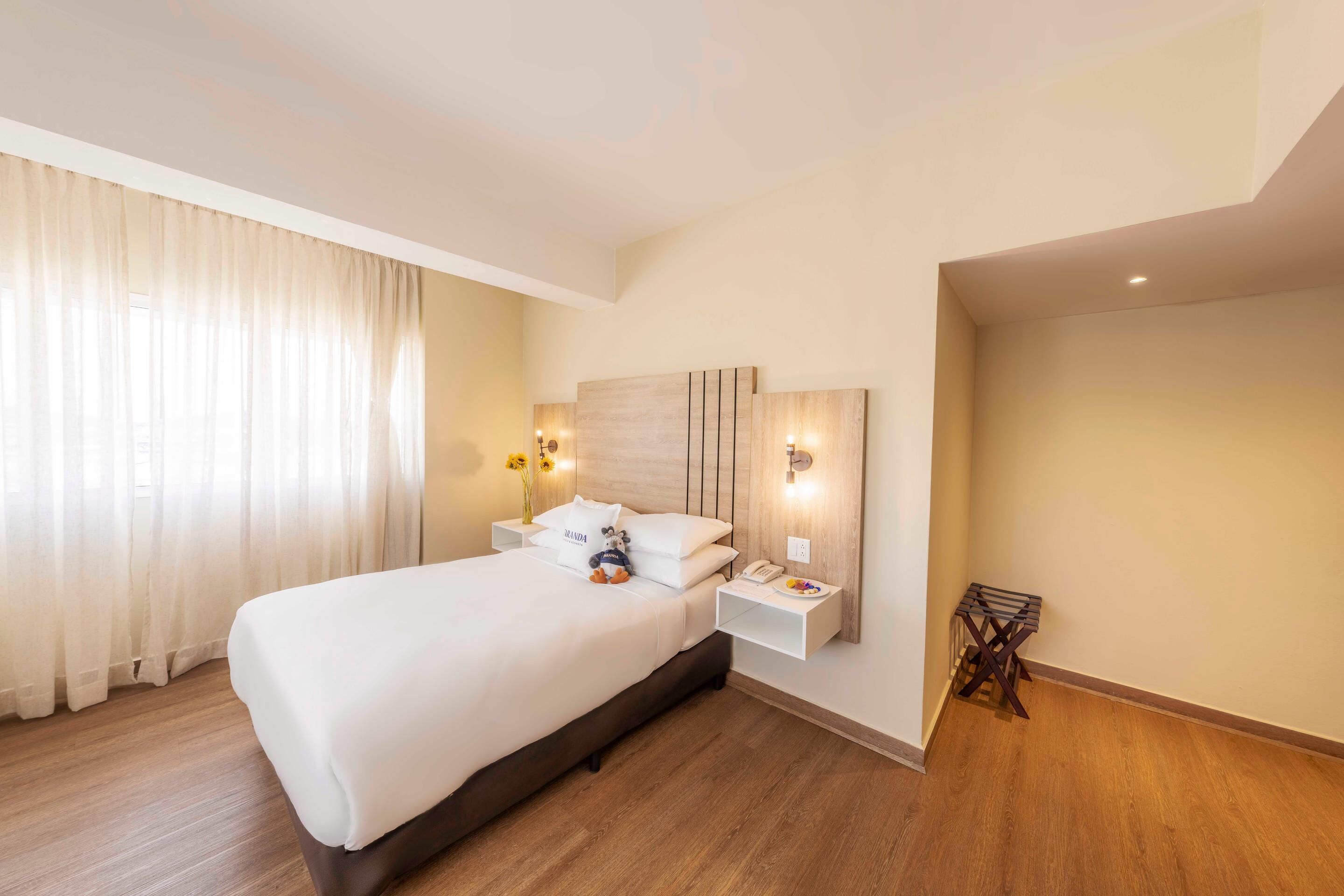 A hotel room with a large bed, a bedside table with a lamp, and a small side table with a phone and a plate of fruit.