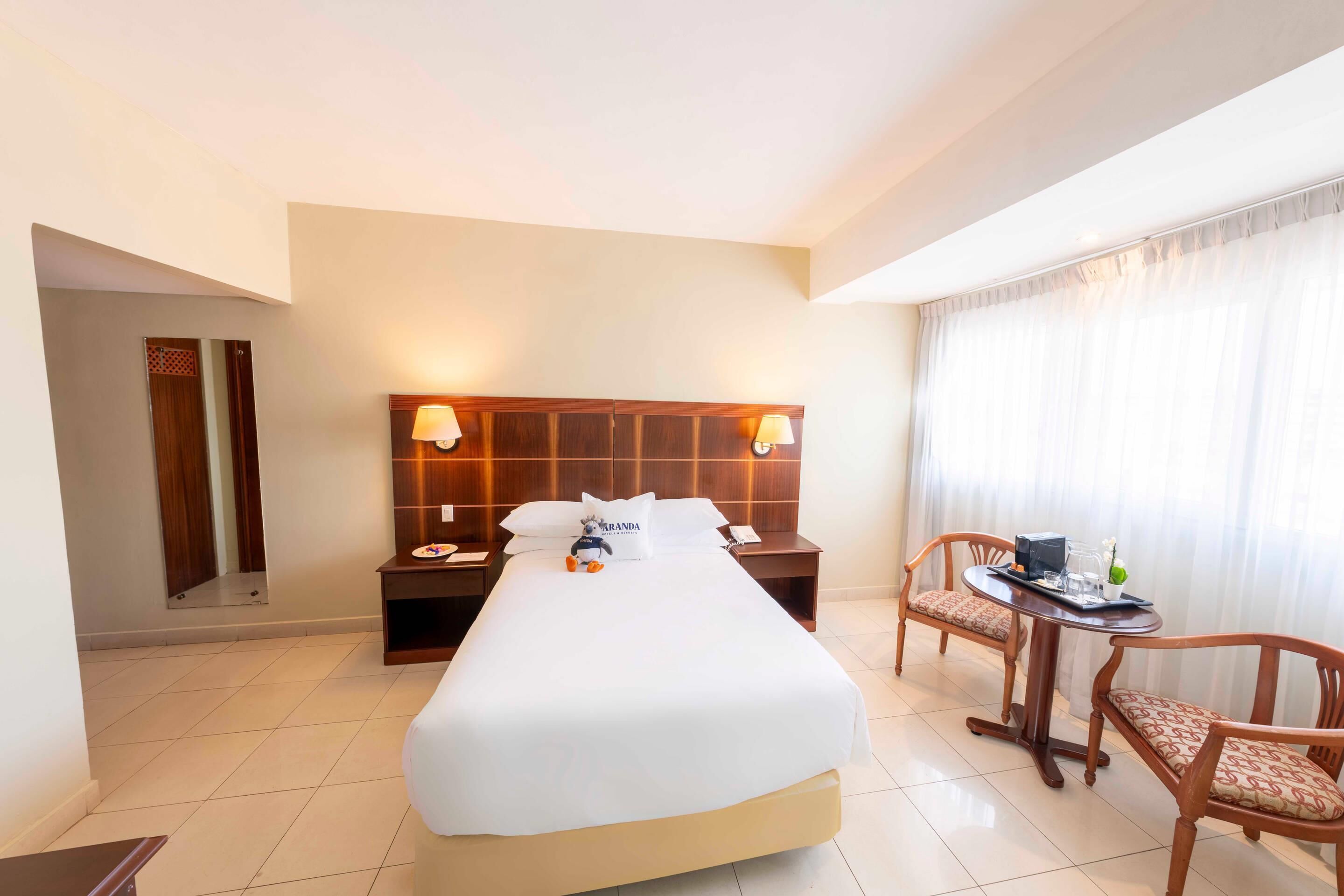 A hotel room with a large bed, a desk with a chair, a small table with a chair, and a window with curtains.