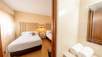 A hotel room with a large bed, a smaller bed, a nightstand with a lamp, a mirror, and a bathroom with a towel rack.