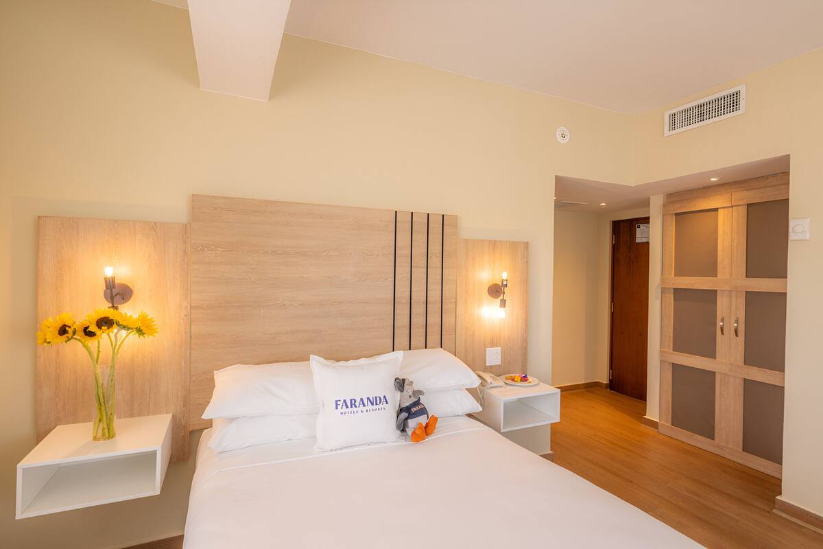 A hotel room with a large bed, a nightstand with a lamp, a wall-mounted light, and a wooden wardrobe.