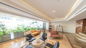 Un lobby espacioso con diseño moderno, que incluye un conjunto de sofás, varias sillas, una mesa de centro y una escalera con barandilla de vidrio.