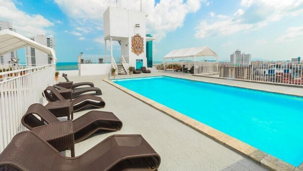 Un área de piscina con sillones de descanso, un edificio blanco con un escudo y una vista despejada del océano.