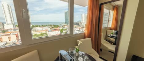 Habitación de hotel con un ventanal que ofrece vistas de la ciudad, una zona de comedor con una mesa preparada para dos personas y un espejo que refleja la habitación.