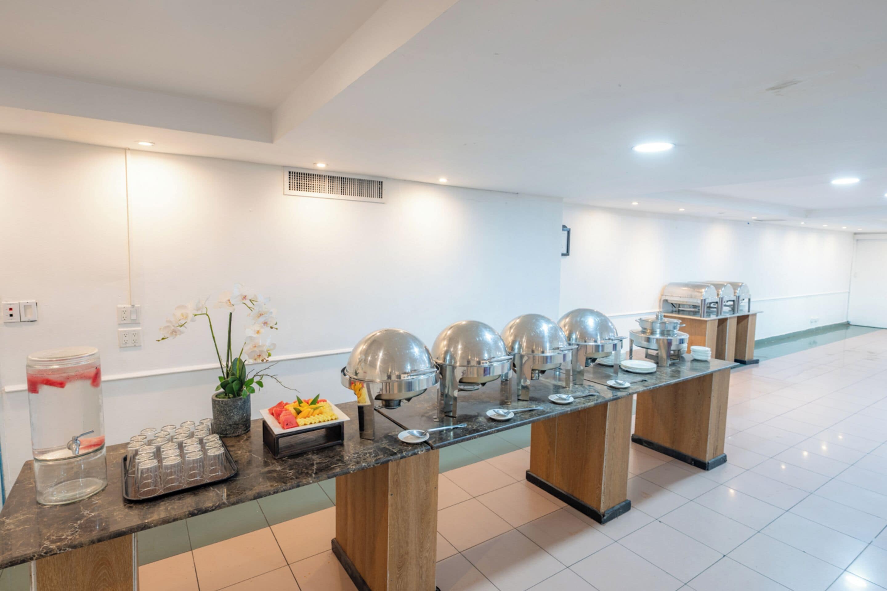 Montaje tipo bufé con varias estaciones de servicio, dispensador de agua y una bandeja de fruta.