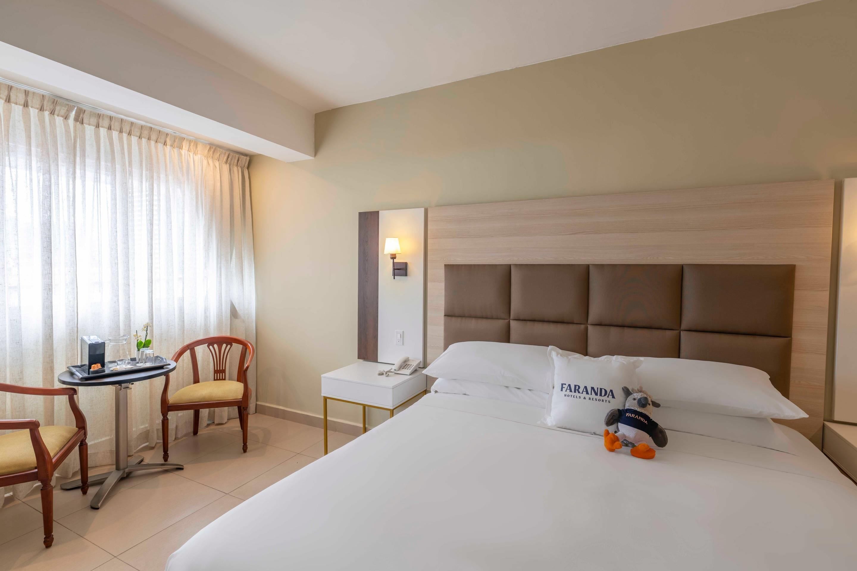 A hotel room with a bed, a desk, a chair, a lamp, and a stuffed toy.