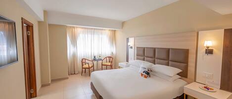 A hotel room with a large bed, a bedside table, a desk, two chairs, a window with curtains, and a wall-mounted lamp.