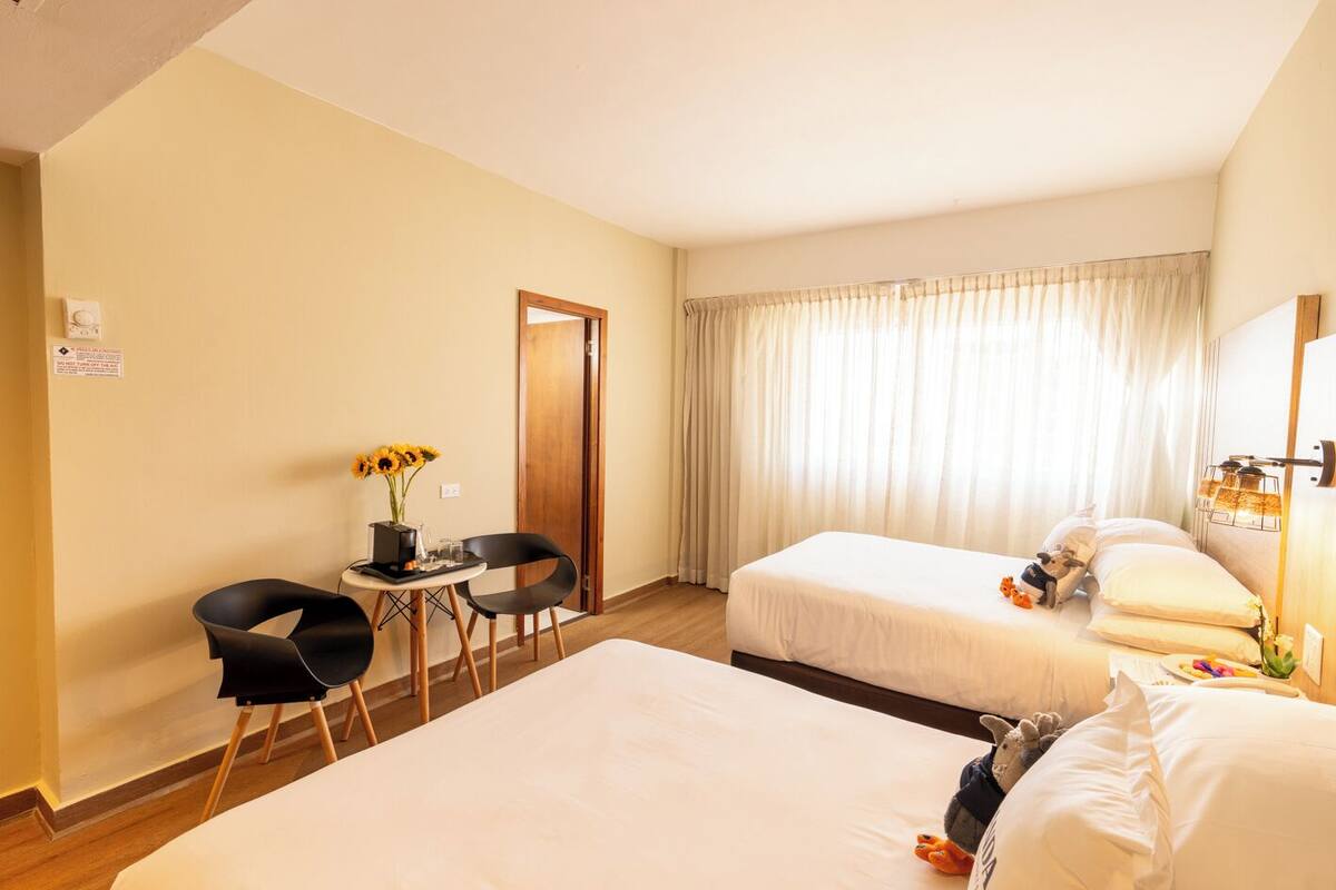 A hotel room with two beds, a small table with chairs, a vase of sunflowers, and a window with curtains.