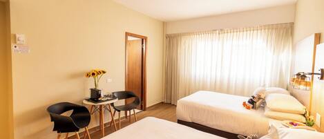A hotel room with two beds, a small table with chairs, a vase of sunflowers, and a window with curtains.