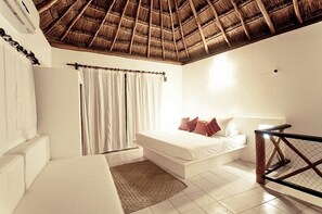 Cozy retreat with a thatched roof, featuring a comfortable bed, seating area, and large window with white curtains.
